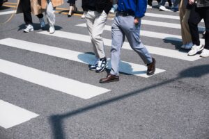 Pedestrians crossing a street at a crosswalk in broad daylight, showing diverse footwear.