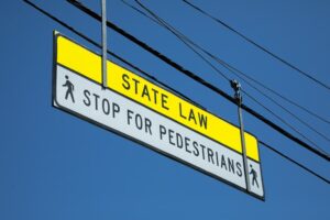 "State Law. Stop For Pedestrian" sign hanging above a crosswalk, with a blue sky background