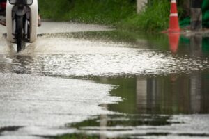 A motorcycle rides through a large puddle on a wet road, splashing water around.