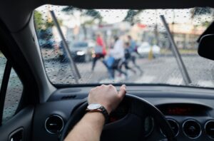 Driver's view through a rainy windshield, with pedestrians crossing a street ahead in blurred focus.