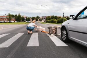 Woman kneeling on a crosswalk, assisting a fallen pedestrian near a car after an accident.