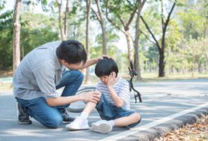 A father comforts his child who fell off a bicycle and injured his knee.